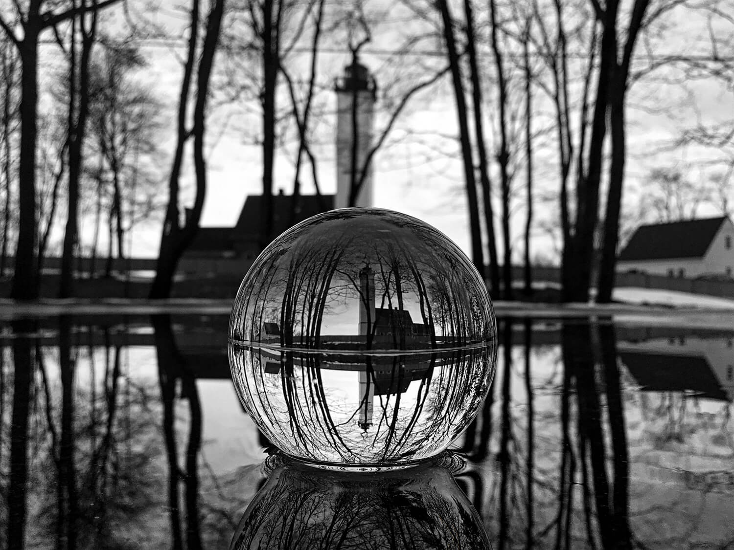 A black and white photograph of a crystal ball reflecting a lighthouse and trees in a reflective surface