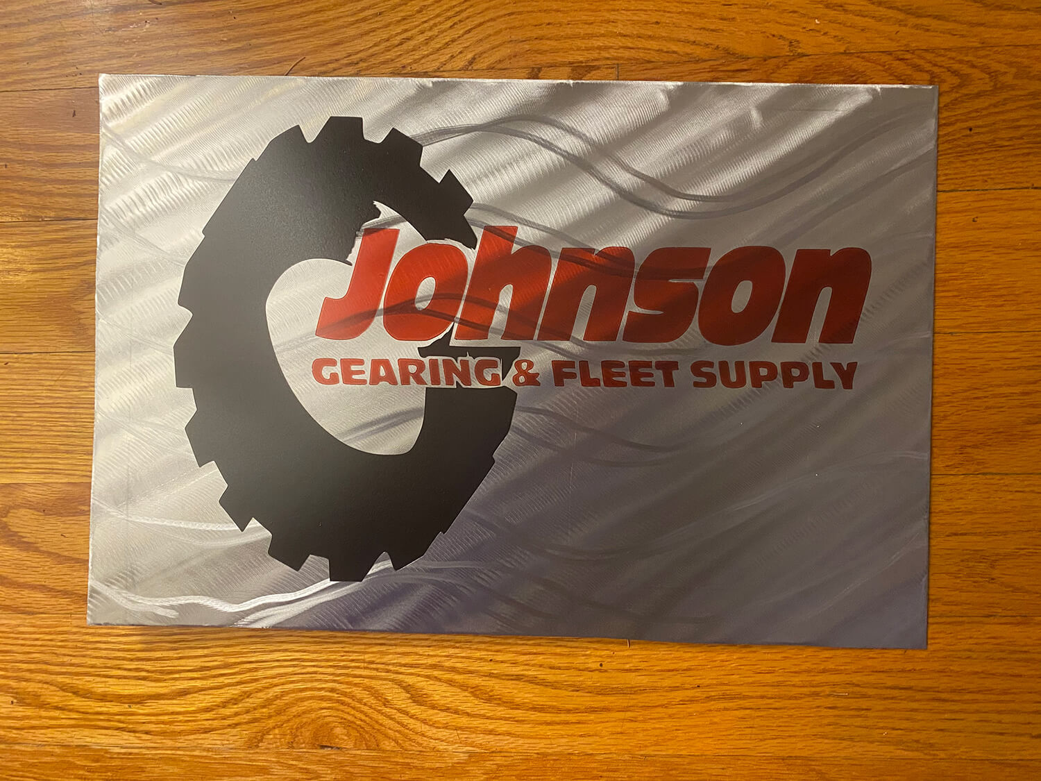Metal sign with the words "Johnson Gearing & Fleet Supply" and a black tire logo over a light grey, wavy background 