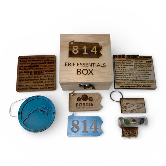A flat lay display of the "814 Erie Essentials Box" surrounded by various Erie, PA themed gifts, including laser-engraved coasters, a Lake Erie keychain, a blue Presque Isle ornament, and a glass vial of genuine Lake Erie beach glass, an 814 magnet and three Erie classic recipes.