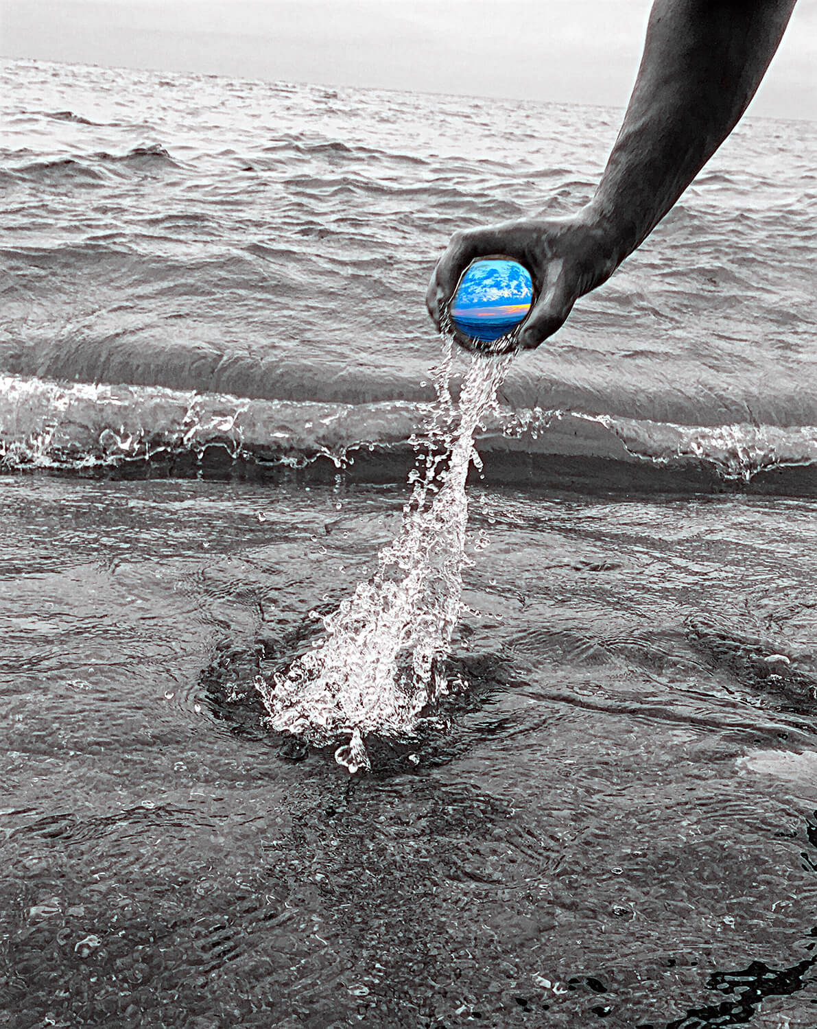 A photograph of a hand holding a small crystal ball above water with a black and white background